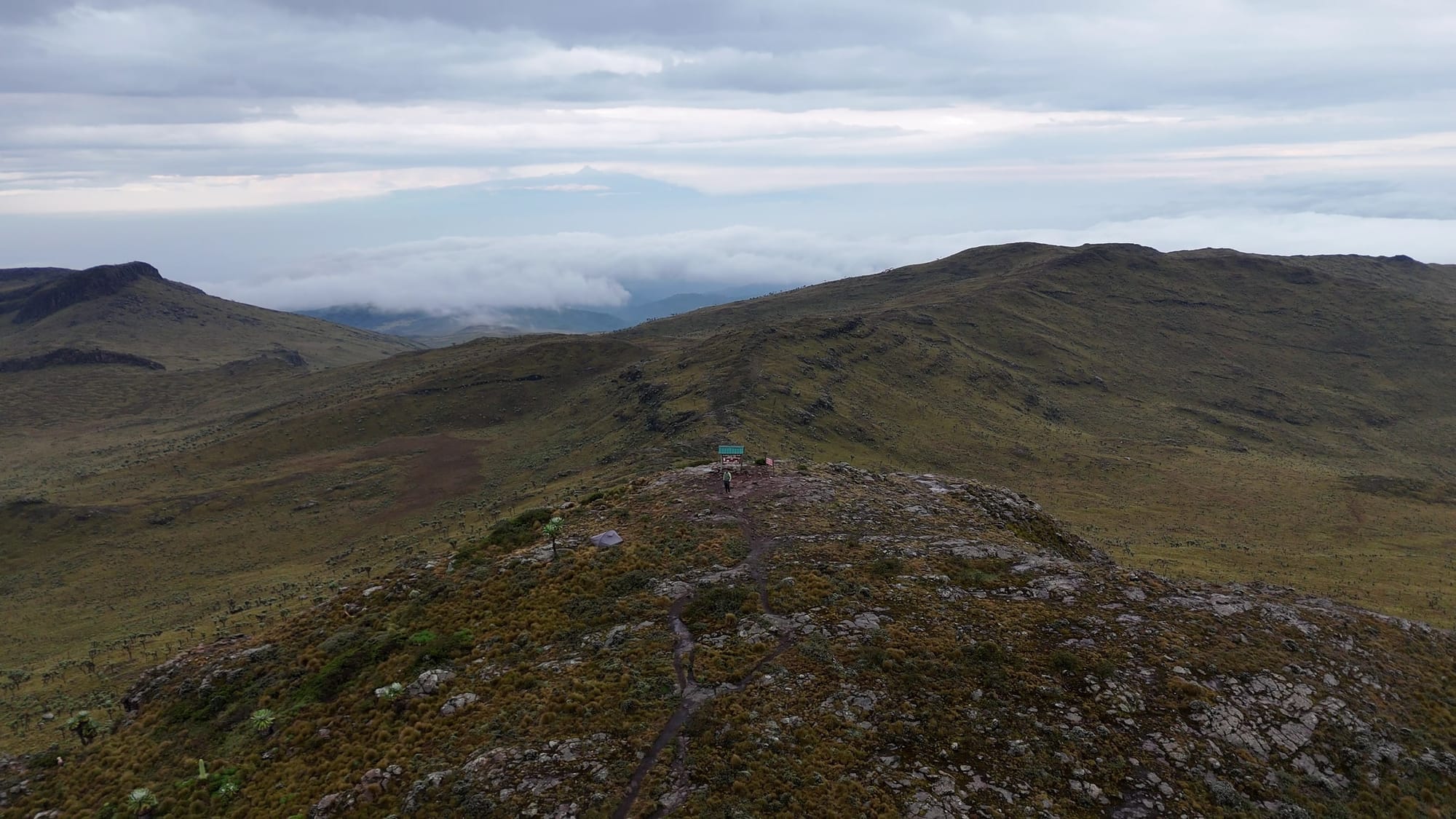 A rocky ridge leads towards the Satima summit marker, surrounded by open hills and low cloud.